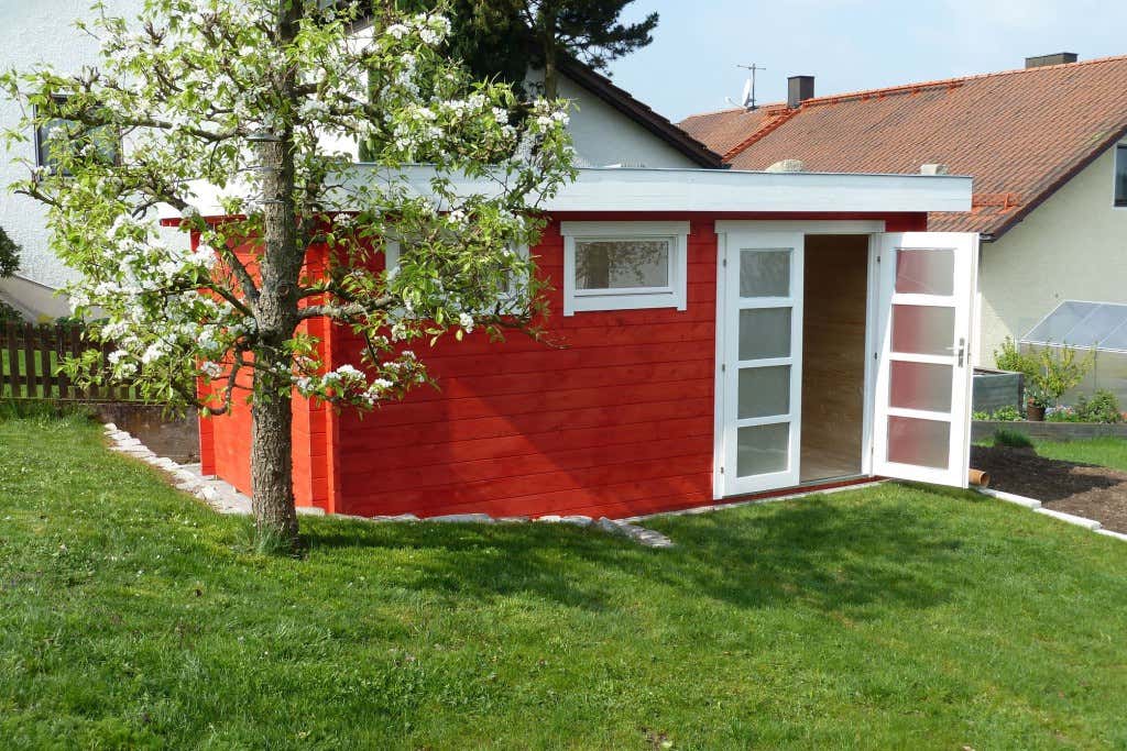 Dieses moderne Flachdach-Gartenhaus Kyara-44 in schwedenrot steht im Schatten eines Obstbaumes. Hier würde sich eine gemütliche Sitzgruppe wunderbar machen, finden Sie nicht?