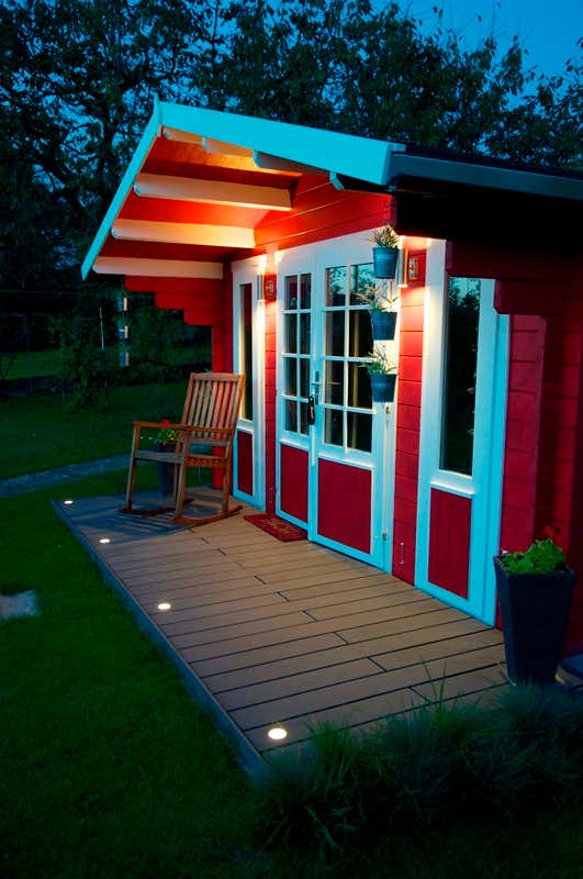 Mit integrierten Lampen im Terrassenboden erstrahlt Ihr schwedenrotes Gartenhaus (Modell Sauerland A-58) in neuem Licht – und in lauen Sommernächten können Sie auf der Terrasse lauschige Abendstunden verbringen.